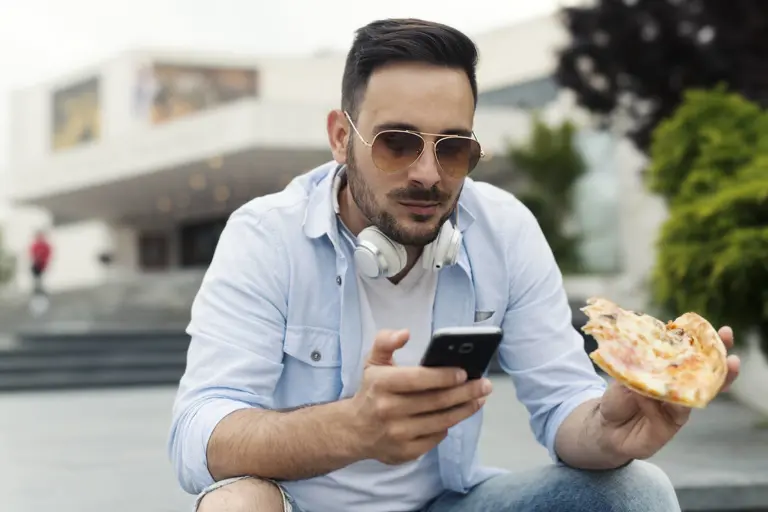 a man using a smartphone and eating a large slice of pizza